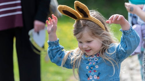 A little girl wearing furry bunny ears and a flowery blue dress smiles with her hands in the air.